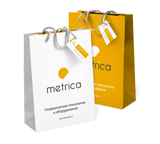 Белый и оранжевый фирменные бумажные пакеты группы компаний «Метрика» с прикрепленными к ручкам корпоративными визитными карточками